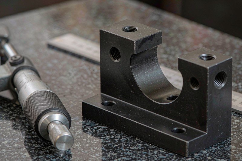 Blackened steel bracket with tapped holes on a granite surface plate beside a micrometer