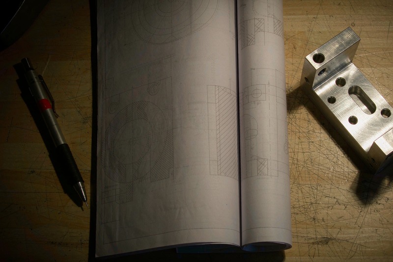 Printed engineering drawing spread on a bench beside a machined aluminum bracket
