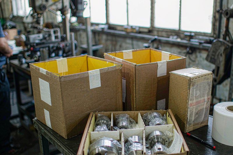 Divided box of machined housings, cartons and a roll of labels on a packing bench