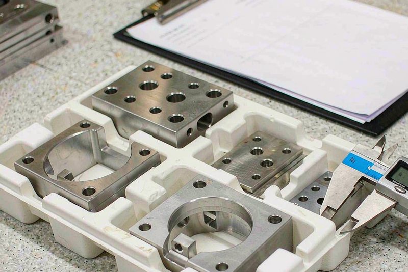 Tray of machined stainless and aluminum parts with a digital caliper on a surface plate